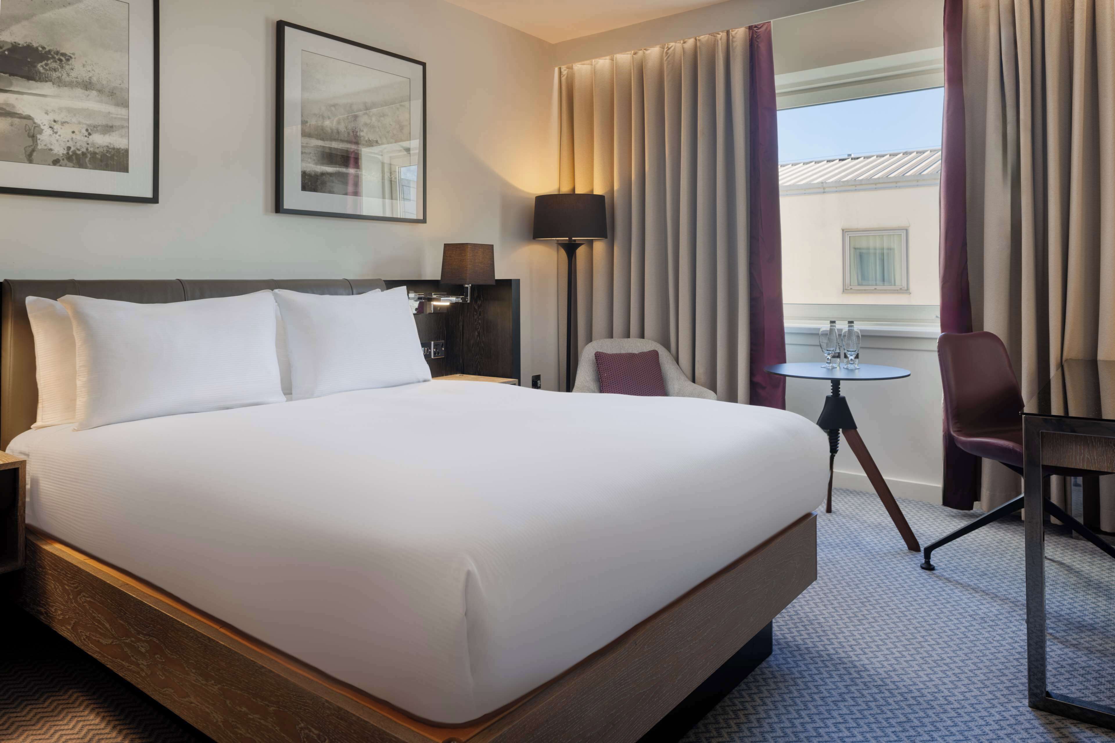 Habitación de hotel con una cama grande, un escritorio con dos sillas, una lámpara y un ventanal con cortinas.