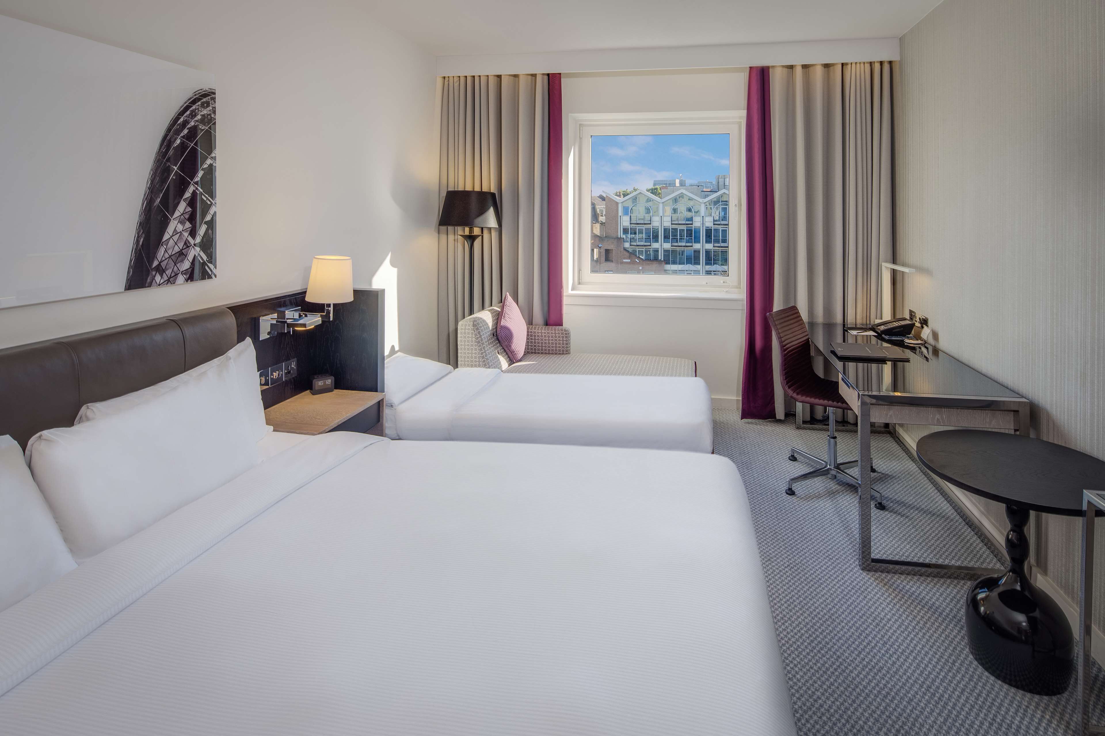 A hotel room with two beds, a desk, a chair, a lamp, and a window with curtains.