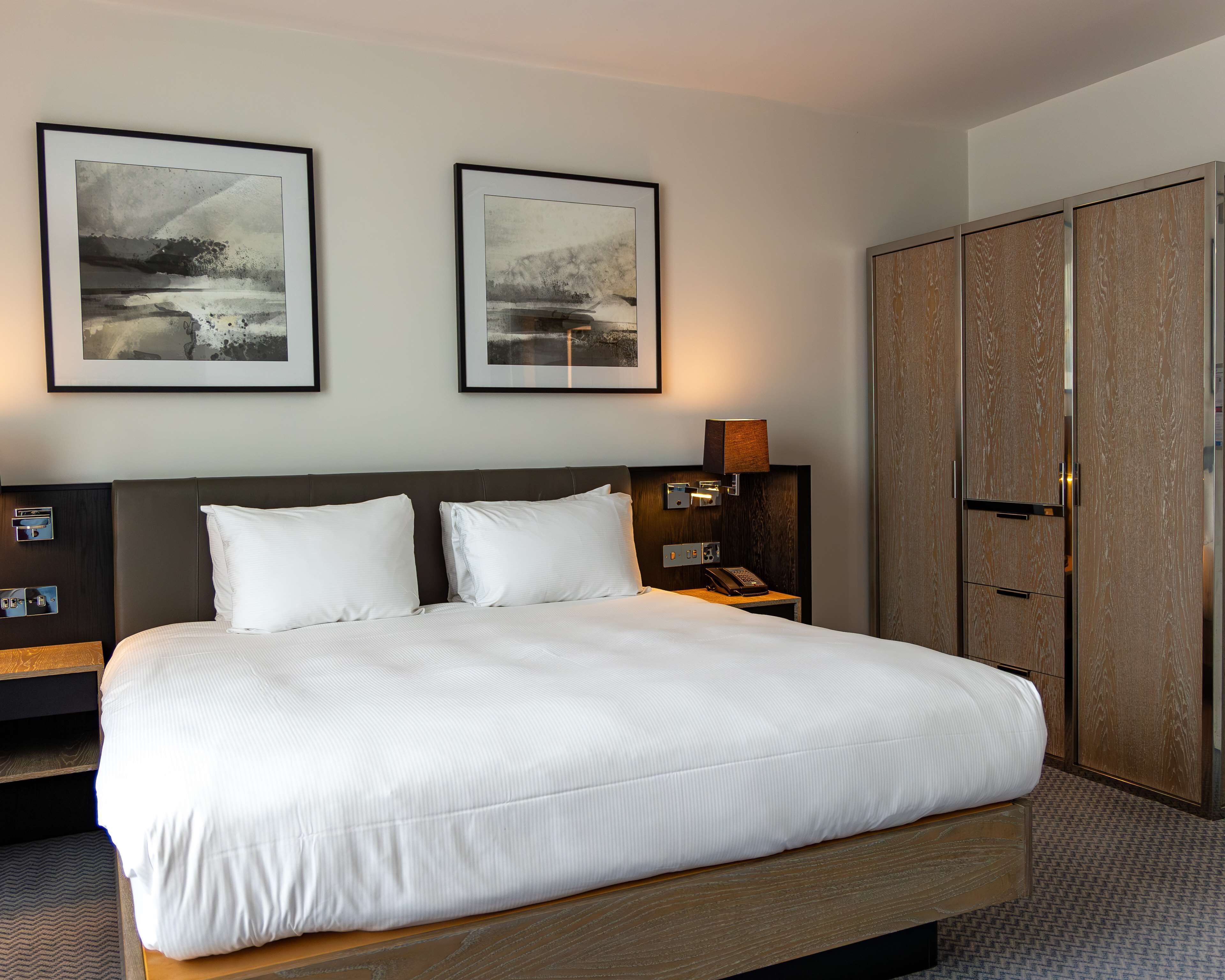 Habitación de hotel con una cama grande, dos cuadros en la pared, una mesita de noche con una lámpara y un armario.