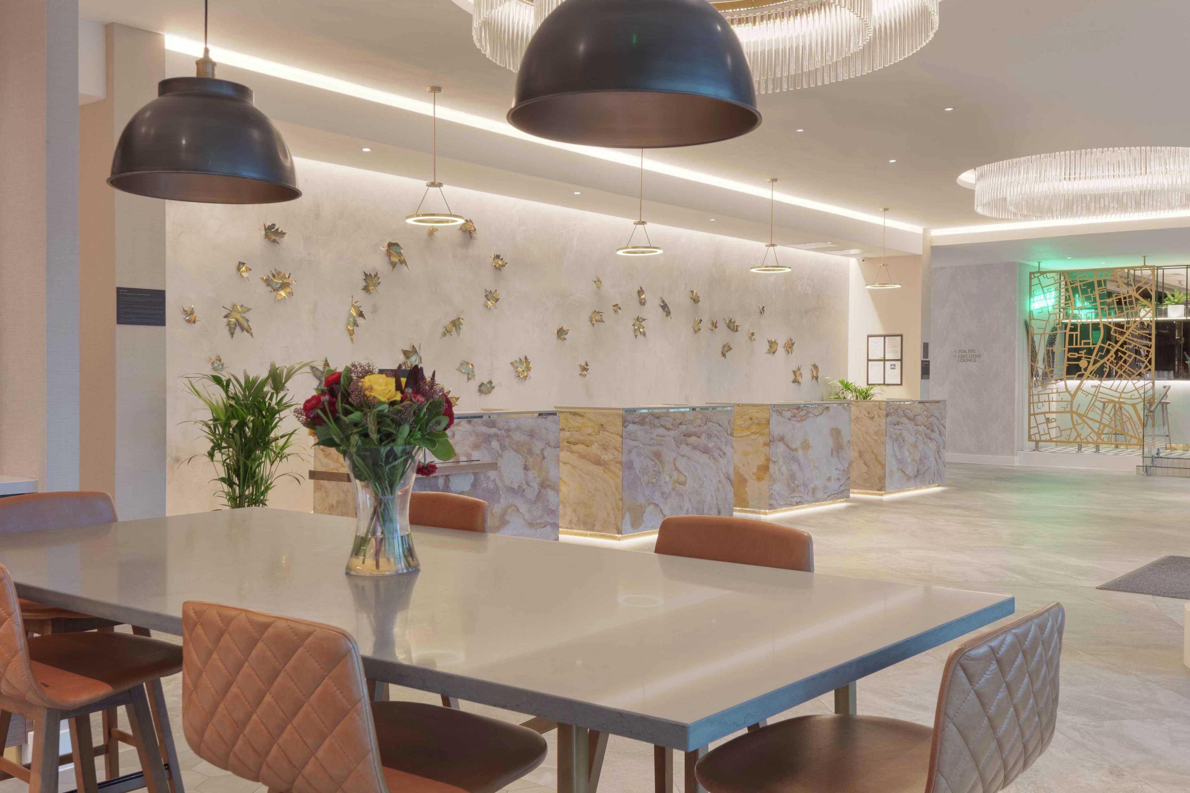 A modern hotel lobby with a marble reception desk, pendant lighting, and a large dining table with cushioned chairs.