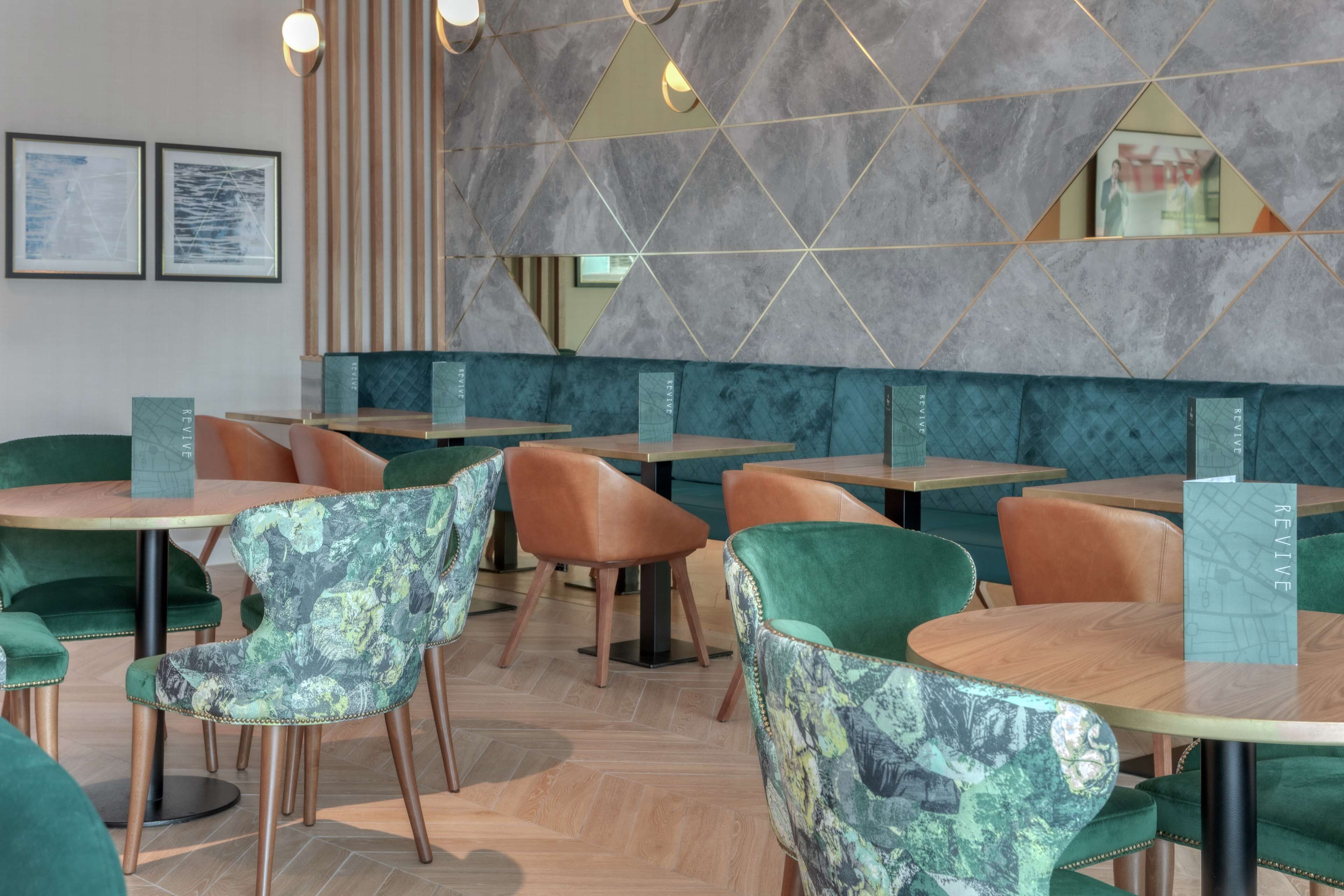 A modern restaurant interior with green upholstered chairs, wooden tables, and a geometric wall design.