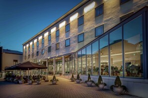 A modern hotel with a glass facade, outdoor seating area, and potted plants. - Hilton London Angel Islington (London)