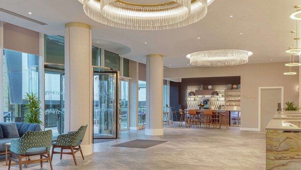 Un vestíbulo amplio con techos altos, grandes columnas y candelabros modernos. Hay zonas de estar con sillones estampados y una zona de bar con taburetes.