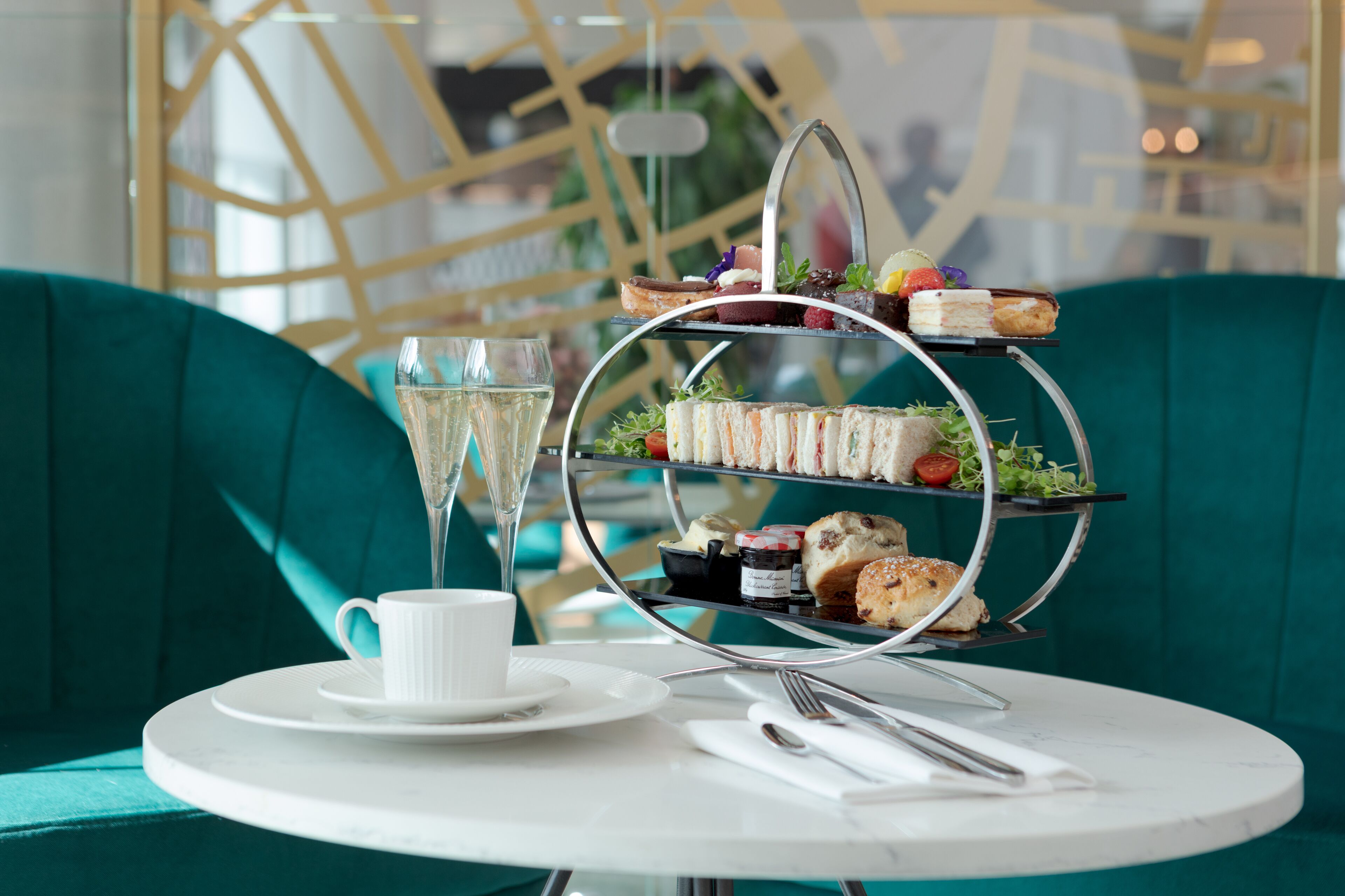 A three-tiered dessert stand with various pastries and sandwiches, a glass of champagne, and a cup on a white table with a green upholstered sofa in the background.