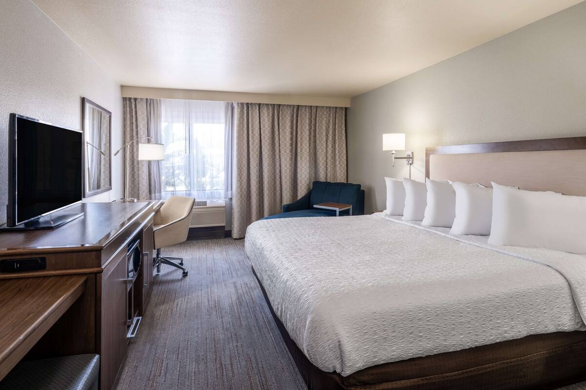 A hotel room with a large bed, a desk with a TV, a chair, and a window with curtains.