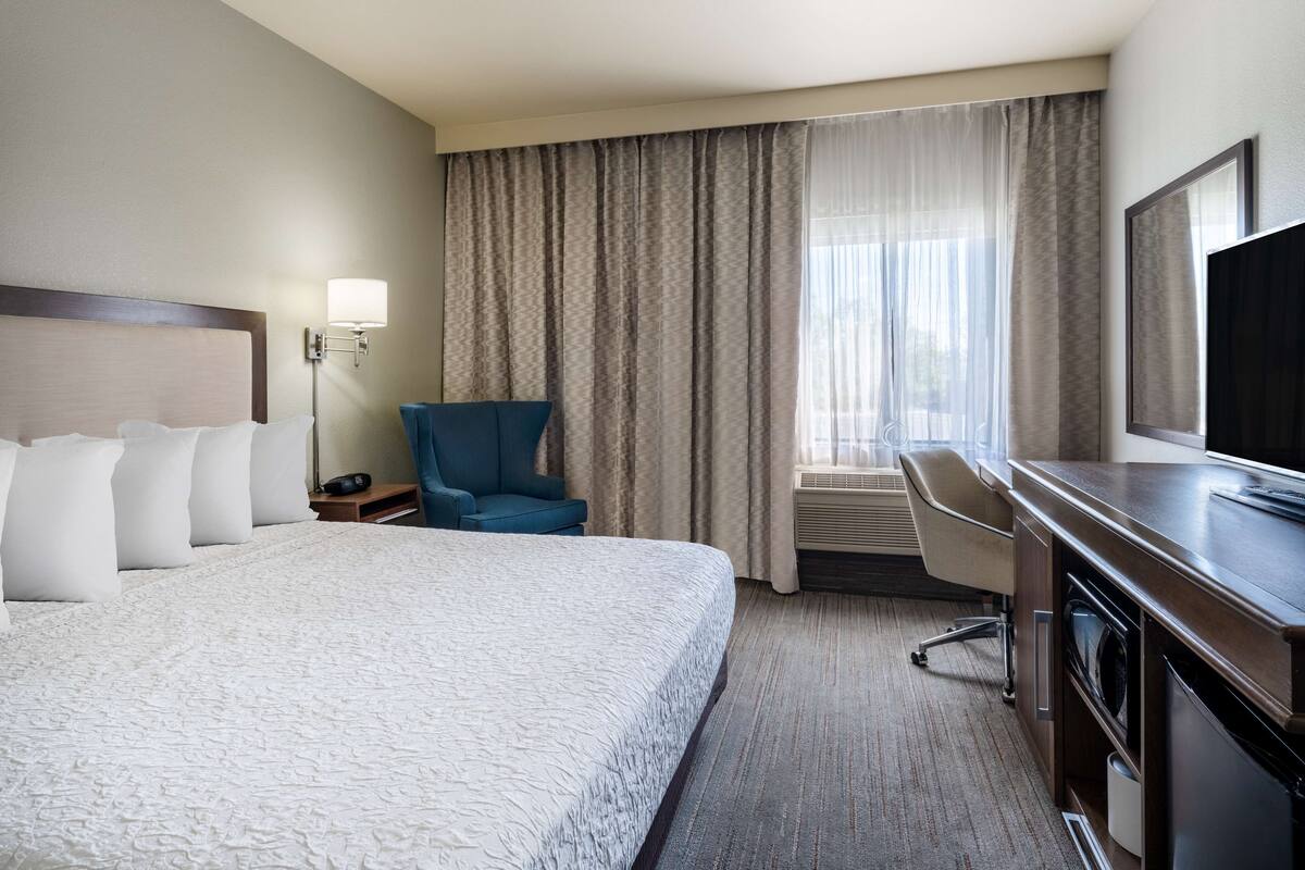 A hotel room with a large bed, a desk, a chair, a TV, and a window with curtains.