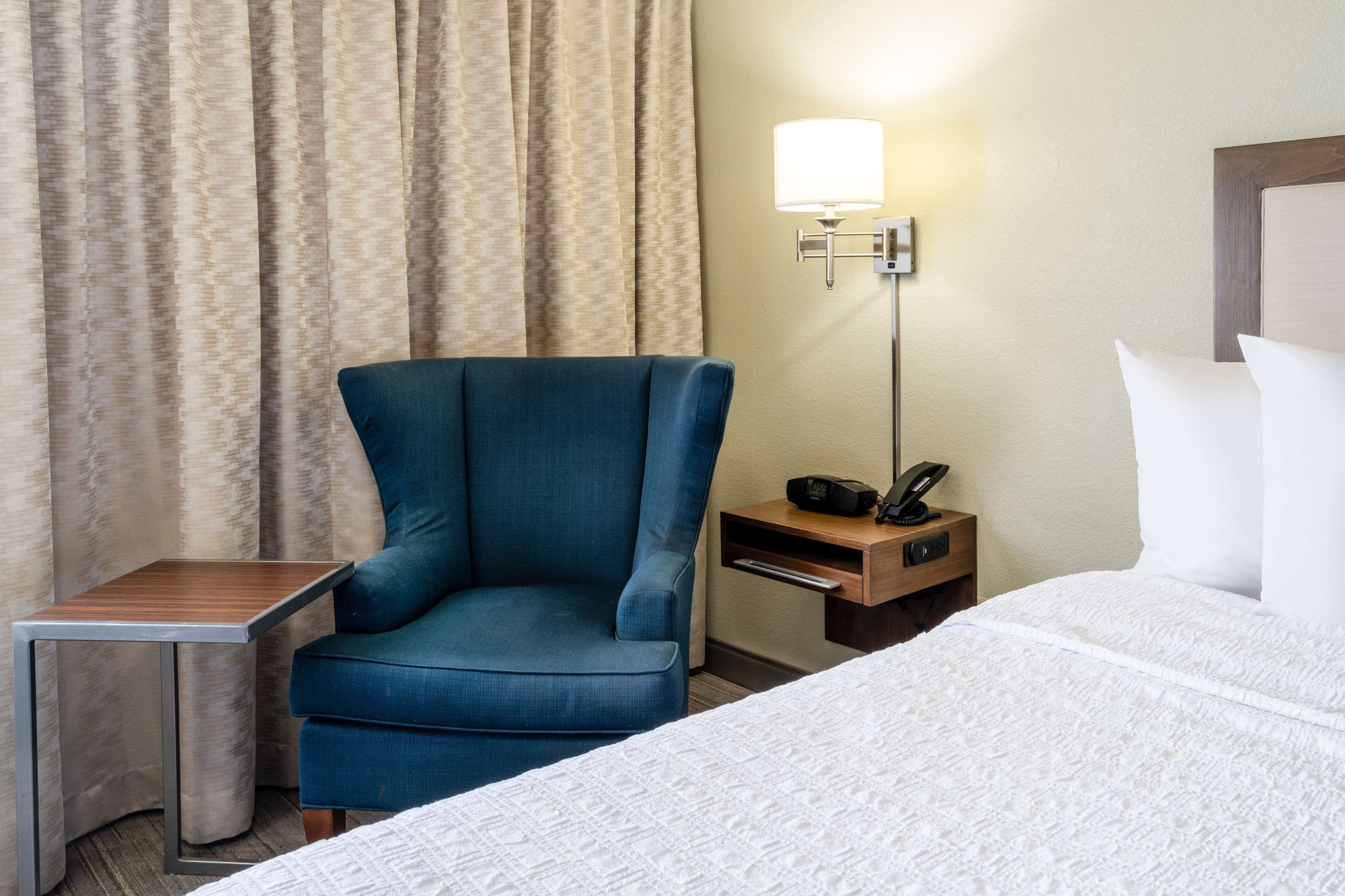 Et hotelværelse med en seng, en blå lænestol, et lille bord, et natbord med en lampe og en telefon.