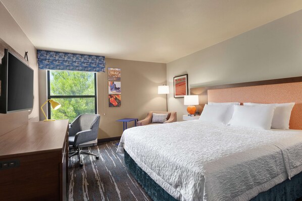 Quarto de hotel com uma cama grande, escrivaninha, cadeira, televisão e janela com vista para árvores.