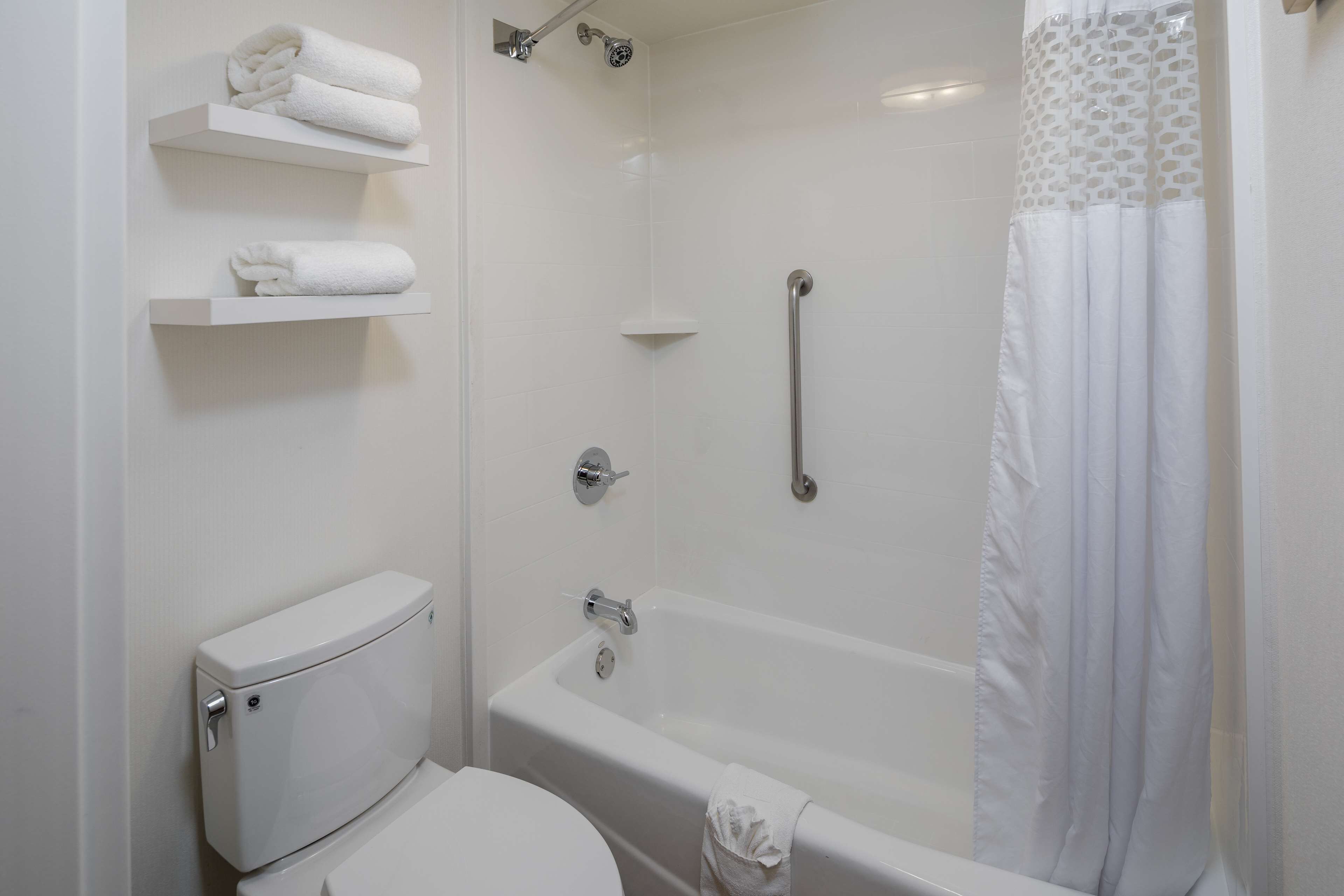 Une salle de bain équipée de toilettes, d’une baignoire et d’une douche. On y trouve des étagères blanches sur lesquelles sont posées des serviettes.