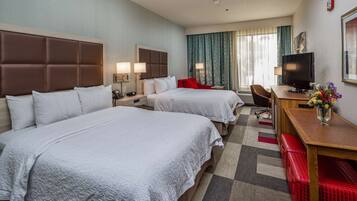 A hotel room with two beds, a desk, a chair, a TV, and a small table with a vase of flowers.