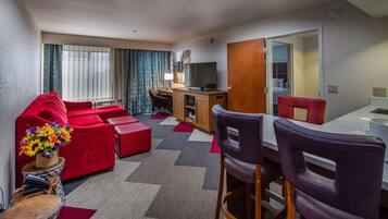 A hotel room with a red sofa, a dining table with chairs, a TV on a wooden cabinet, and a window with curtains.