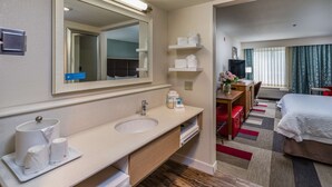 A hotel room with a double bed, a desk with a chair, a large mirror, a sink with a vanity, and a small bathroom area. - Hampton Inn San Francisco Airport (South San Francisco)