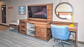 A hotel room with a wooden desk, a television, a blue office chair, a round mirror, and a framed picture on the wall.