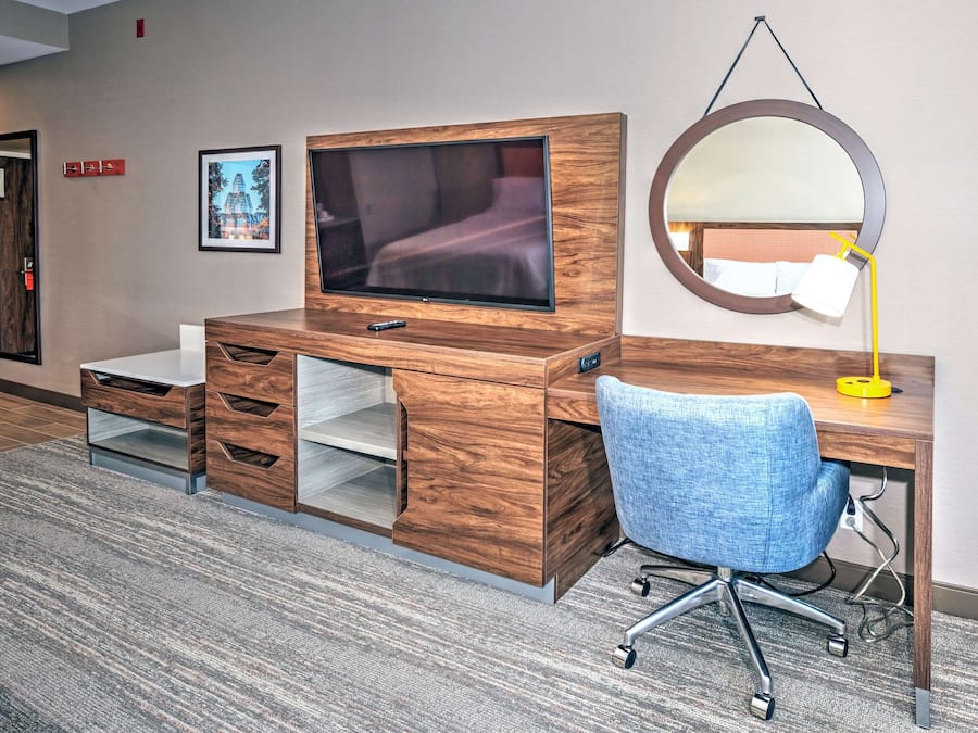 A hotel room with a wooden desk, a television, a blue office chair, a round mirror, and a framed picture on the wall.