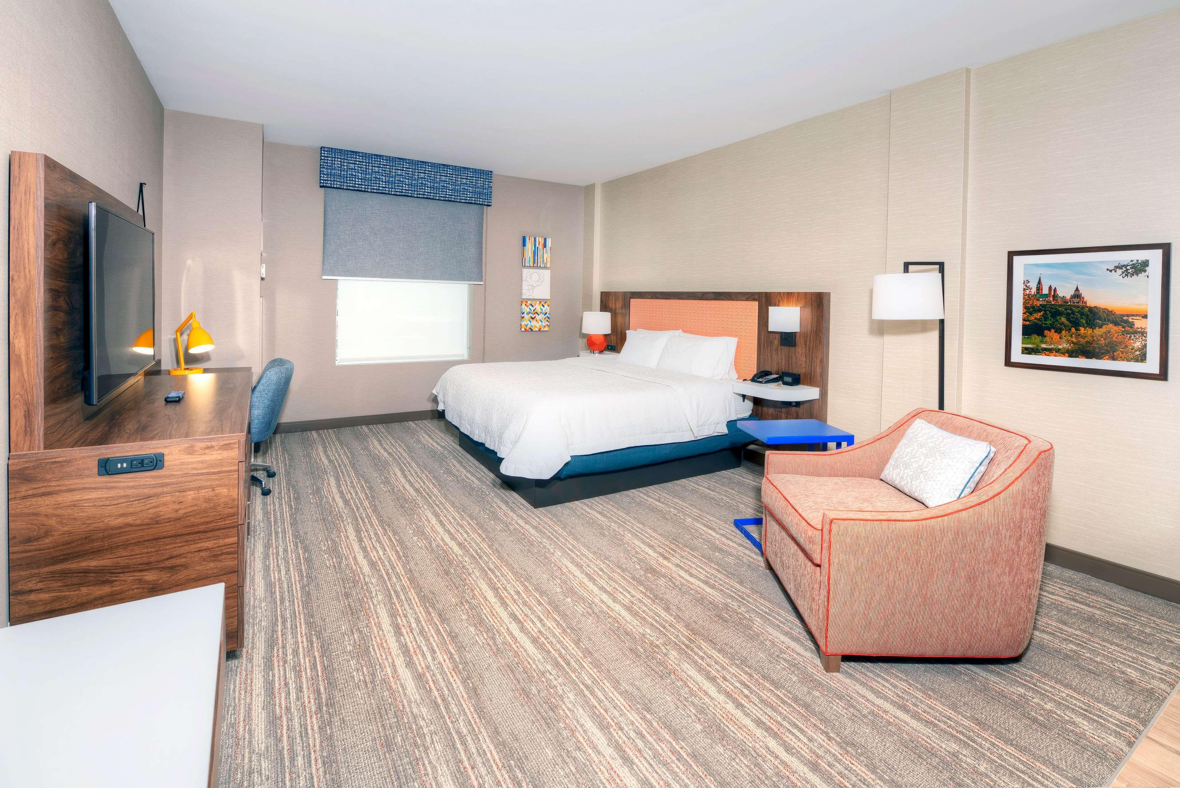A hotel room with a bed, a desk with a TV, a chair, a sofa, a lamp, and a picture on the wall.