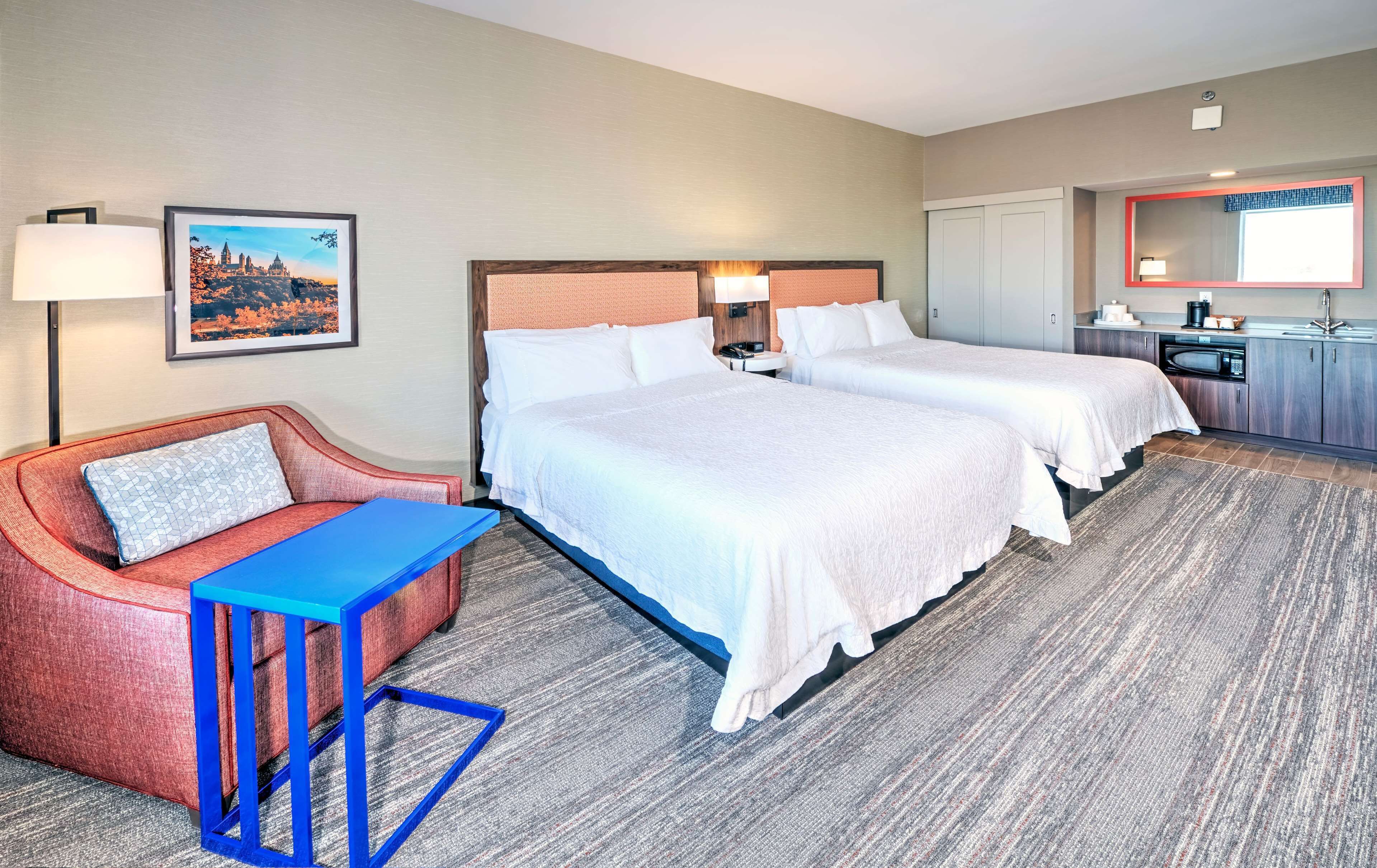 A hotel room with two beds, a desk, a chair, a small table, a lamp, a painting, a mirror, and a kitchenette.