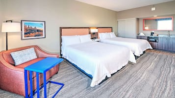Habitación de hotel con dos camas, un escritorio, una silla, una mesita, una lámpara, un cuadro, un espejo y una kitchenette.