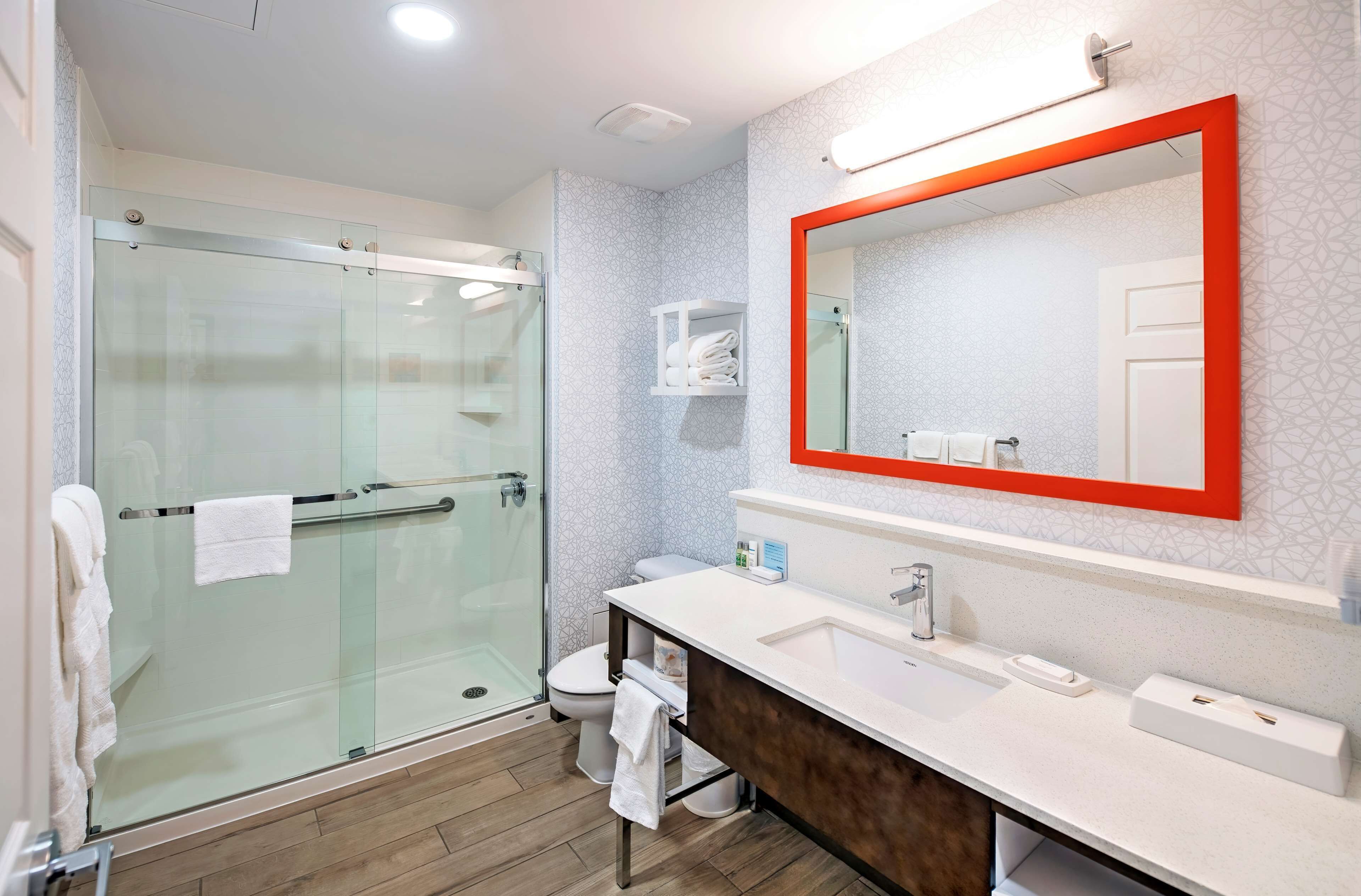 Un baño moderno con ducha de vidrio, un lavamanos blanco con tocador de madera y un espejo grande con marco rojo.