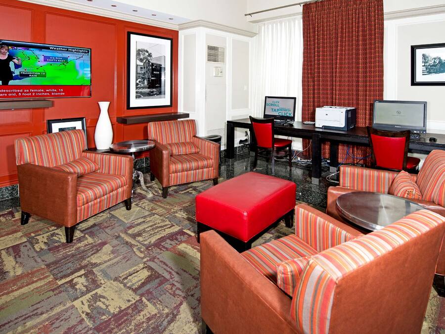 Un vestíbulo de hotel con sofás a rayas, un reposapiés rojo, un televisor mostrando datos del tiempo y cuadros en las paredes.