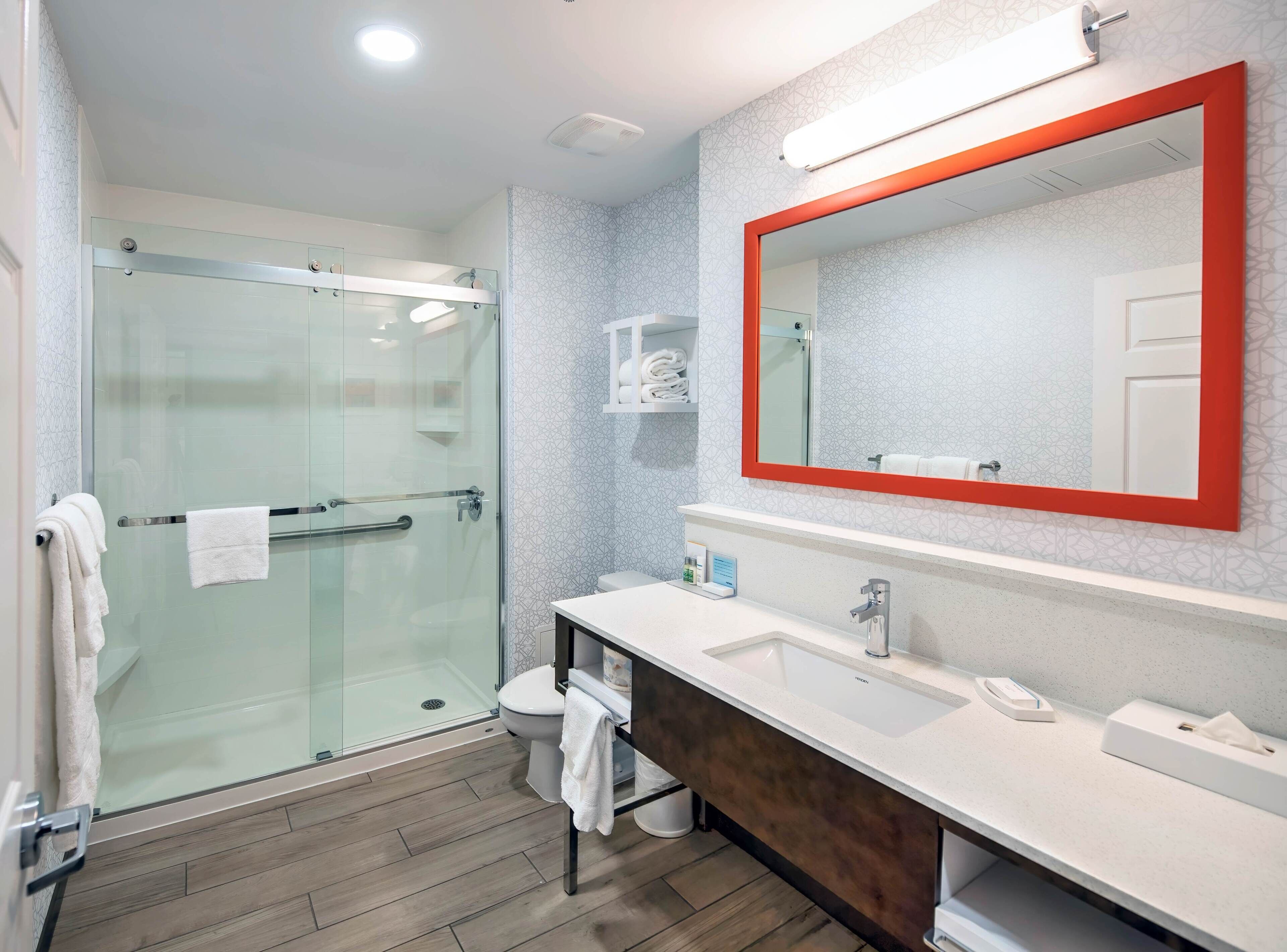 A modern bathroom with a glass shower enclosure, a white sink with a wooden vanity, and a large mirror with a red frame.