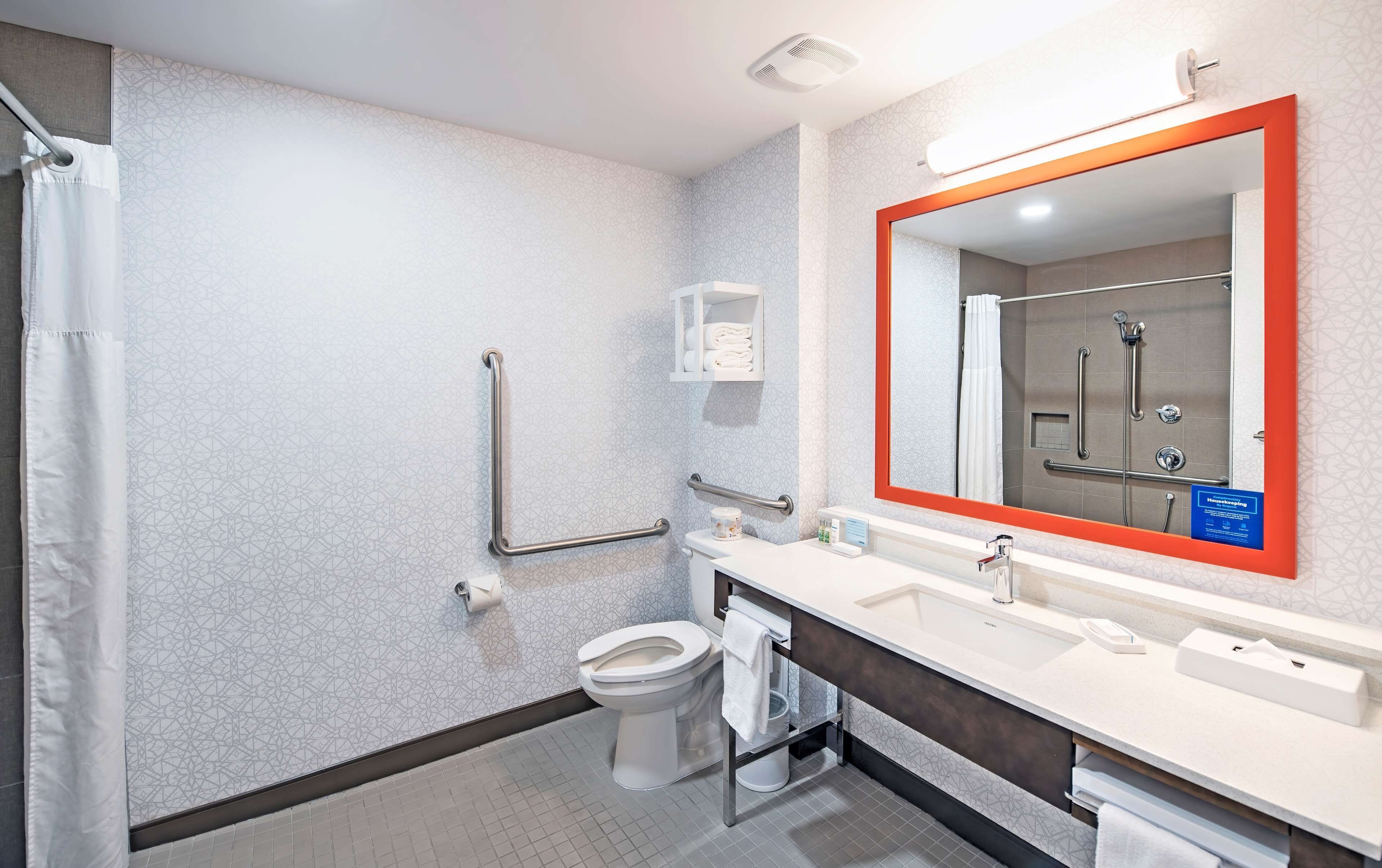 Ein modernes Badezimmer mit Dusche, Toilette und Waschbecken. Es beinhaltet eine Handstange, einen Handtuchhalter und einen Spiegel mit rotem Rahmen.