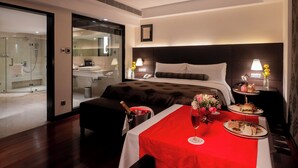 A hotel room with a bed, a bedside table with a lamp, a small table with a red tablecloth and food, and a glass-enclosed shower area. - The LaLiT New Delhi (New Delhi)