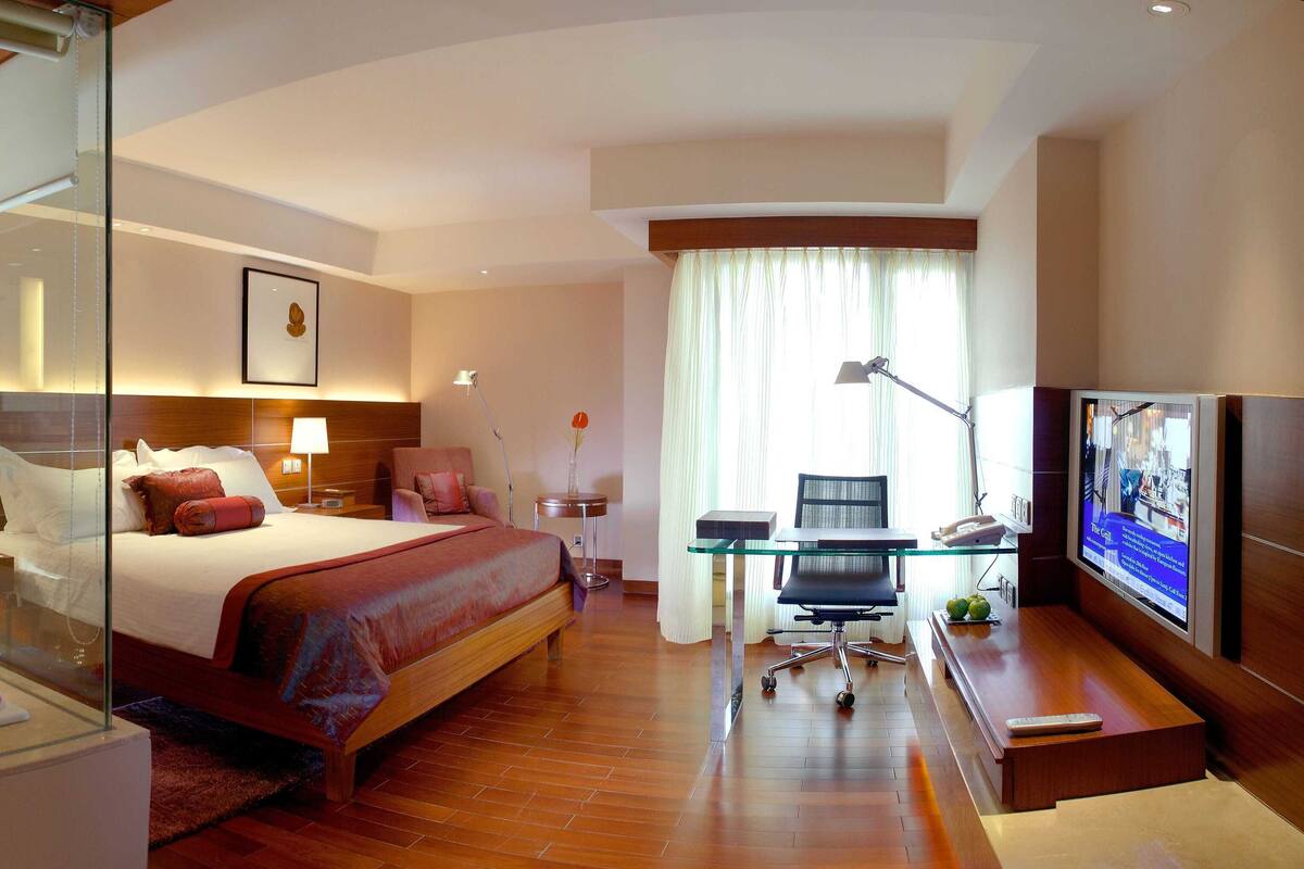 Ein modernes Hotelzimmer mit einem großen Bett, einer gläsernen Dusche, einem Schreibtisch mit Stuhl, einem Fernseher und einem großen Fenster mit Vorhängen.