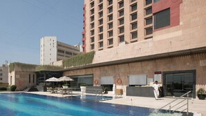 A swimming pool with lounge chairs and umbrellas, a building with a red facade, and a clear sky. - The LaLiT New Delhi (New Delhi)