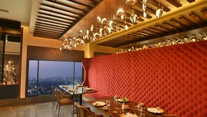 A dining area with a red upholstered booth, a glass table, and a city view through large windows. - The LaLiT New Delhi (New Delhi)