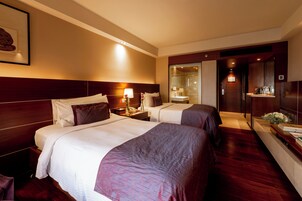 A hotel room with two beds, a nightstand, a lamp, a mirror, and a bathroom visible through a glass door.
