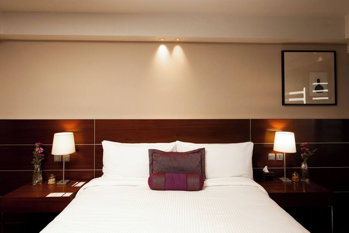 Ein Hotelzimmer mit einem ordentlich bezogenen Bett, zwei Nachttischen mit Lampen, einem Beistelltisch mit einer Vase und einem kleinen Dekorationsgegenstand sowie einem gerahmten Bild an der Wand.
