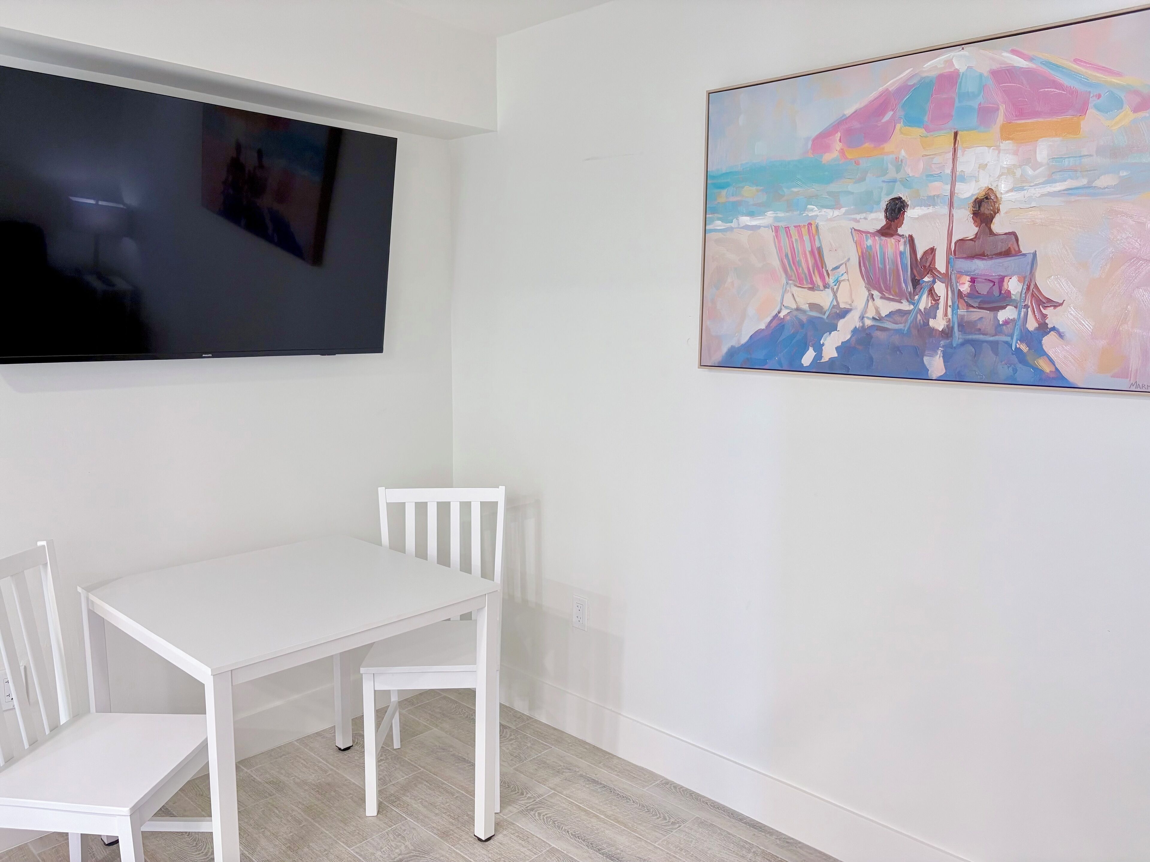Een kamer met een witte tafel en stoelen, een flatscreen televisie aan de muur en een kleurrijk schilderij van een strandgezicht.