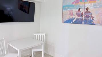 A room with a white table and chairs, a flat-screen TV mounted on the wall, and a colorful beach scene painting on the wall.