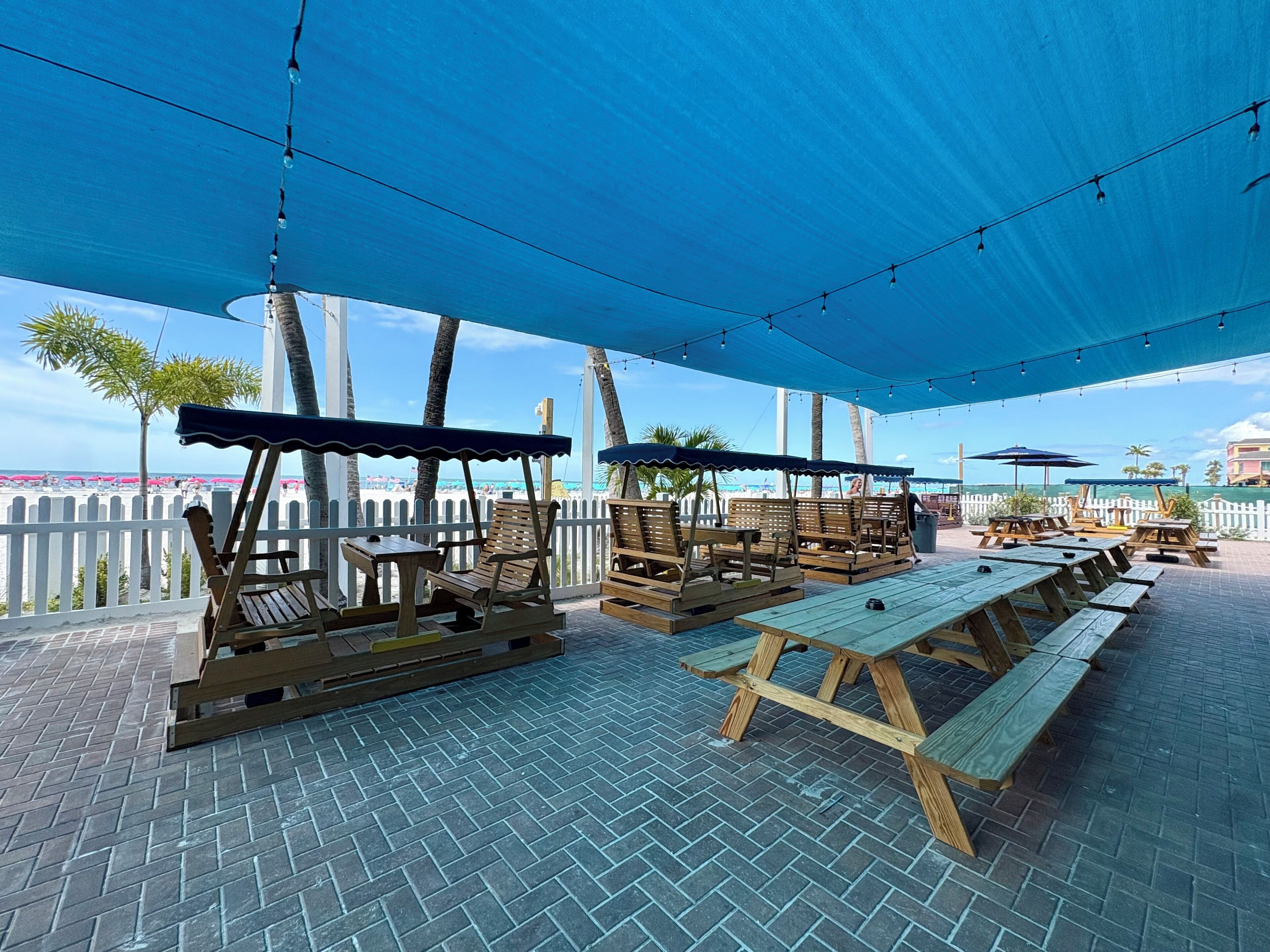 An outdoor patio with wooden benches and picnic tables, a white picket fence, and string lights.