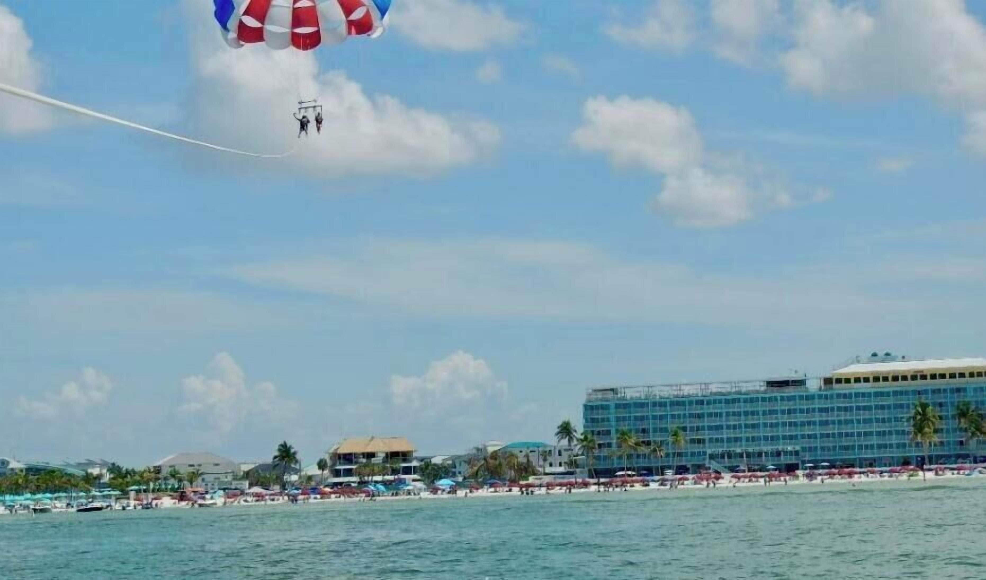 A beach resort with a large hotel building, a sandy beach, and a parasail in the sky.
