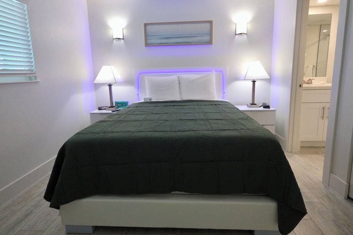 A modern bedroom with a bed, bedside lamps, a painting above the bed, and a bathroom visible through an open door.