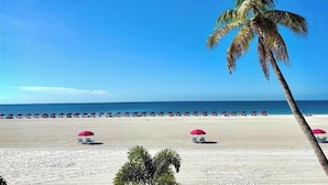 Une plage bordée de rangées de parasols et de chaises longues, de palmiers et d’un ciel bleu azur.