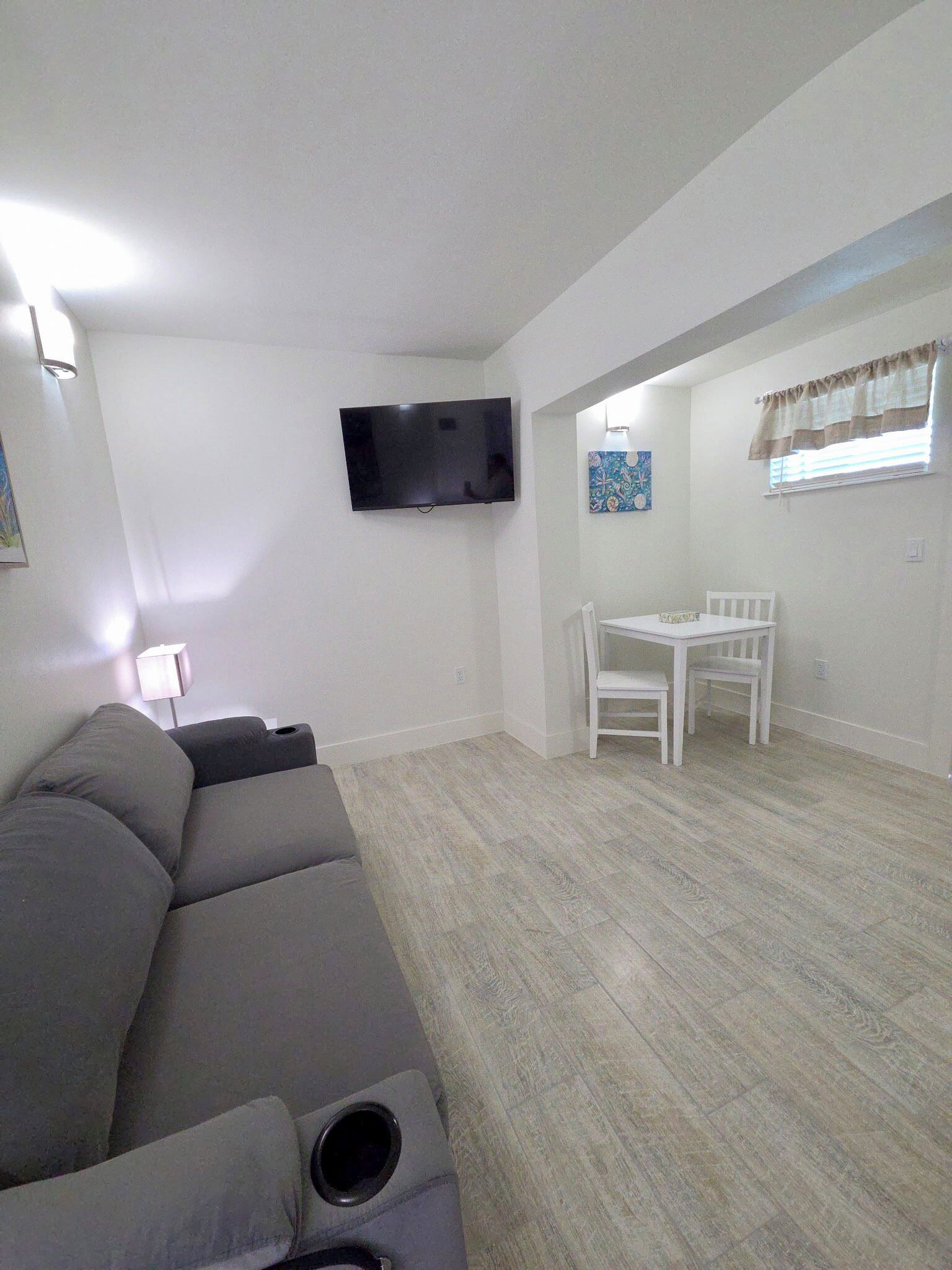 Un salón con un sofá gris, un televisor de pantalla plana montado en la pared, una mesita con sillas y un ventanal con cortinas.