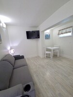 Un salón con un sofá gris, un televisor de pantalla plana montado en la pared, una mesita con sillas y un ventanal con cortinas.