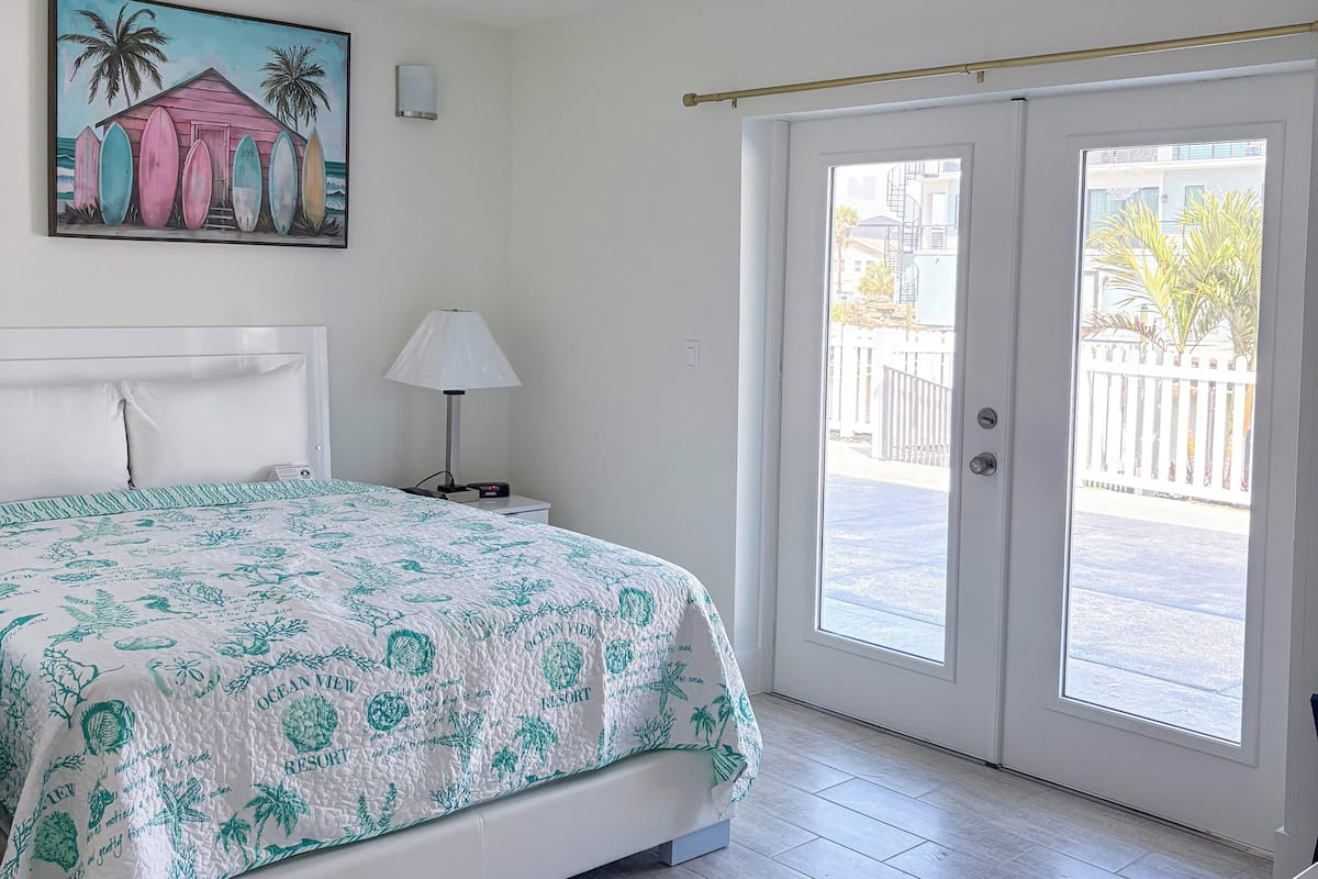A bedroom with a bed, a nightstand, a lamp, a painting, and a sliding glass door leading to an outdoor area.