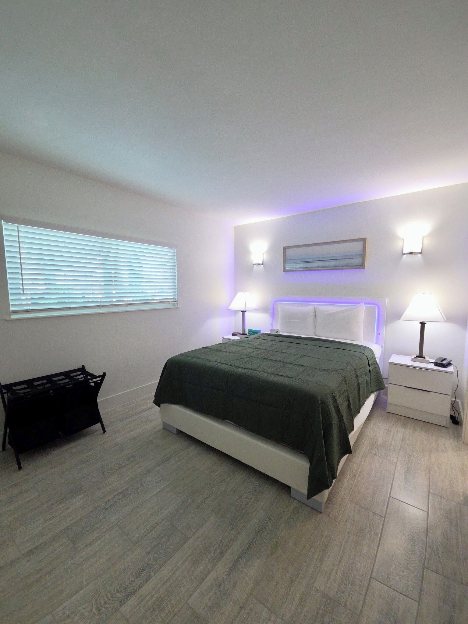 A modern bedroom with a bed, bedside tables, a nightstand, a lamp, a wall-mounted light, a window with blinds, and a painting.