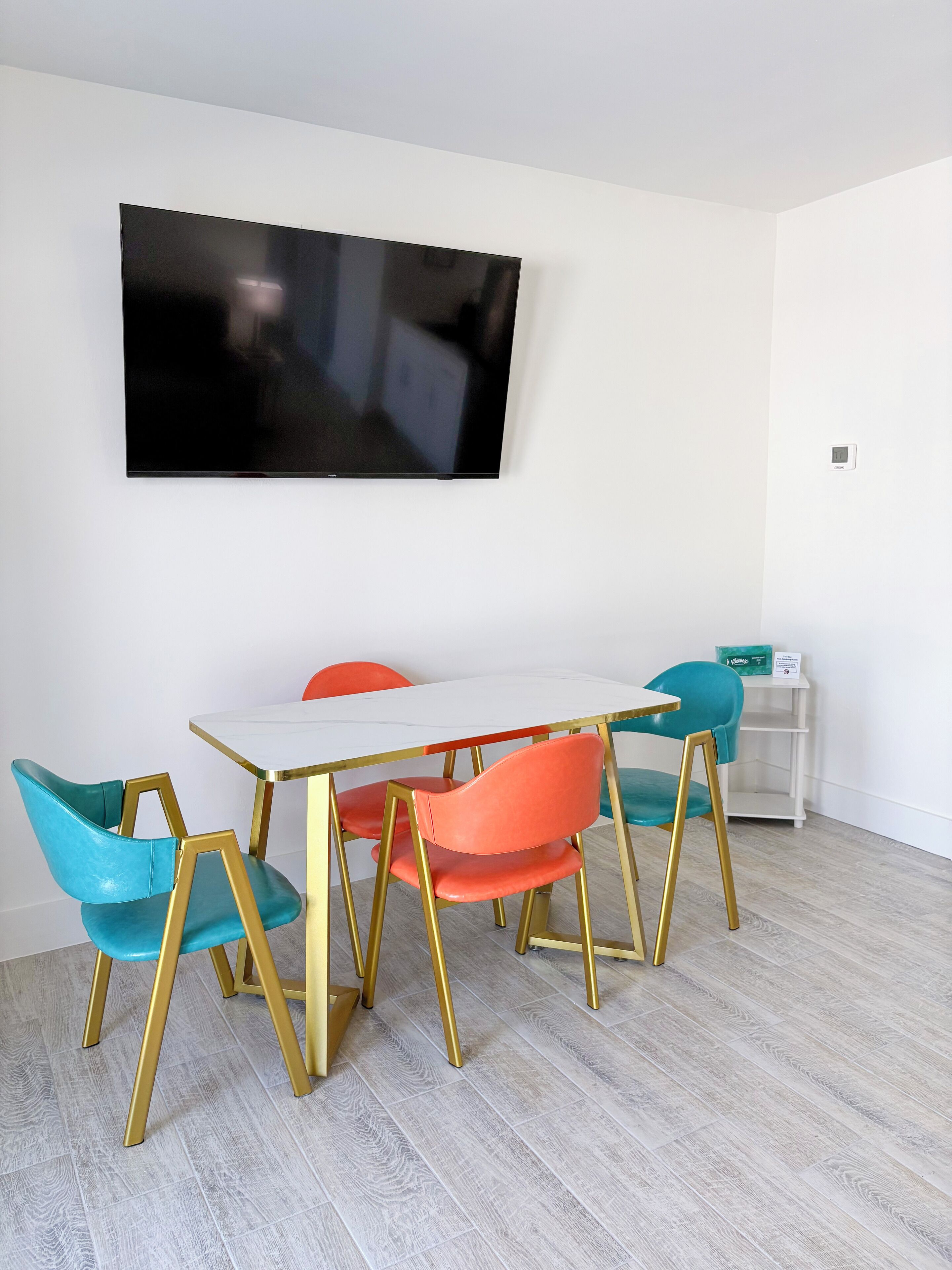 Un comedor moderno con una mesa y sillas blancas de patas doradas, un televisor de pantalla plana montado en la pared y un pequeño armario blanco con un estante.