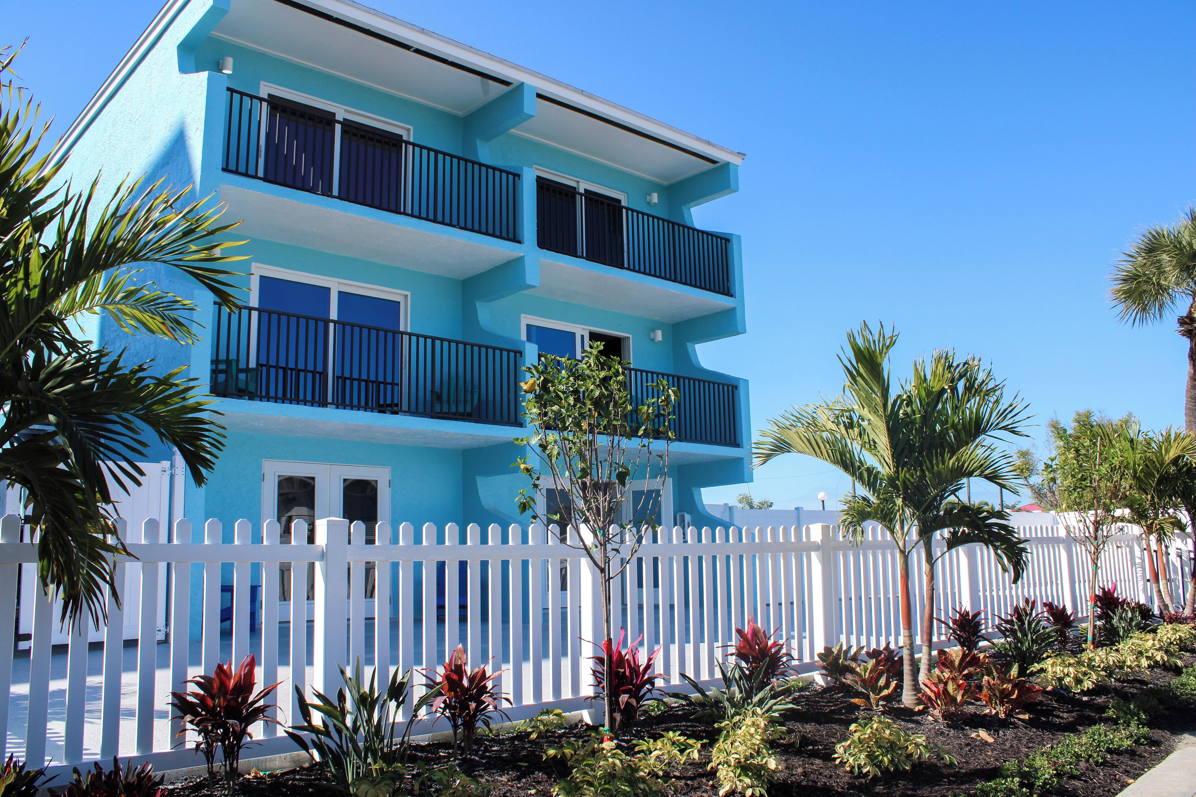 A multi-story building with balconies, a white picket fence, and tropical plants.
