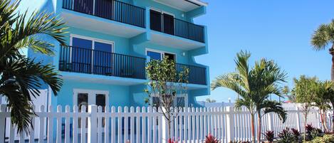 A multi-story building with balconies, a white picket fence, and tropical plants.