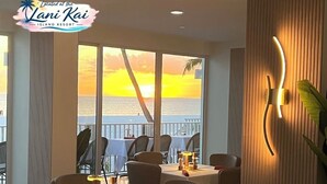 Un espace repas avec des tables préparées pour deux personnes, de grandes fenêtres offrant une vue sur le coucher de soleil sur l’océan et un luminaire mural moderne.