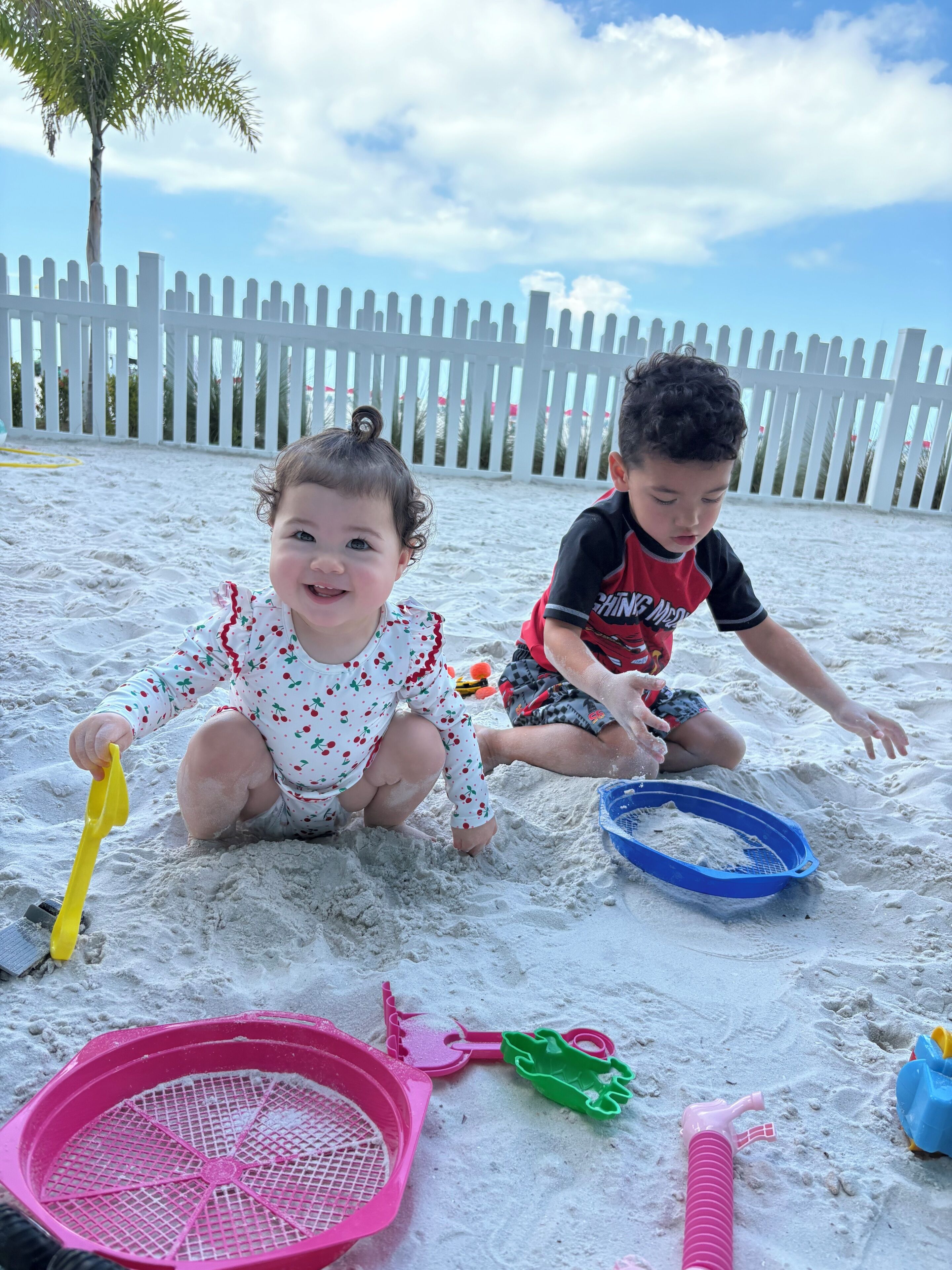 Dos niños jugando en la arena con juguetes de playa, una cerca blanca y un cielo despejado al fondo.