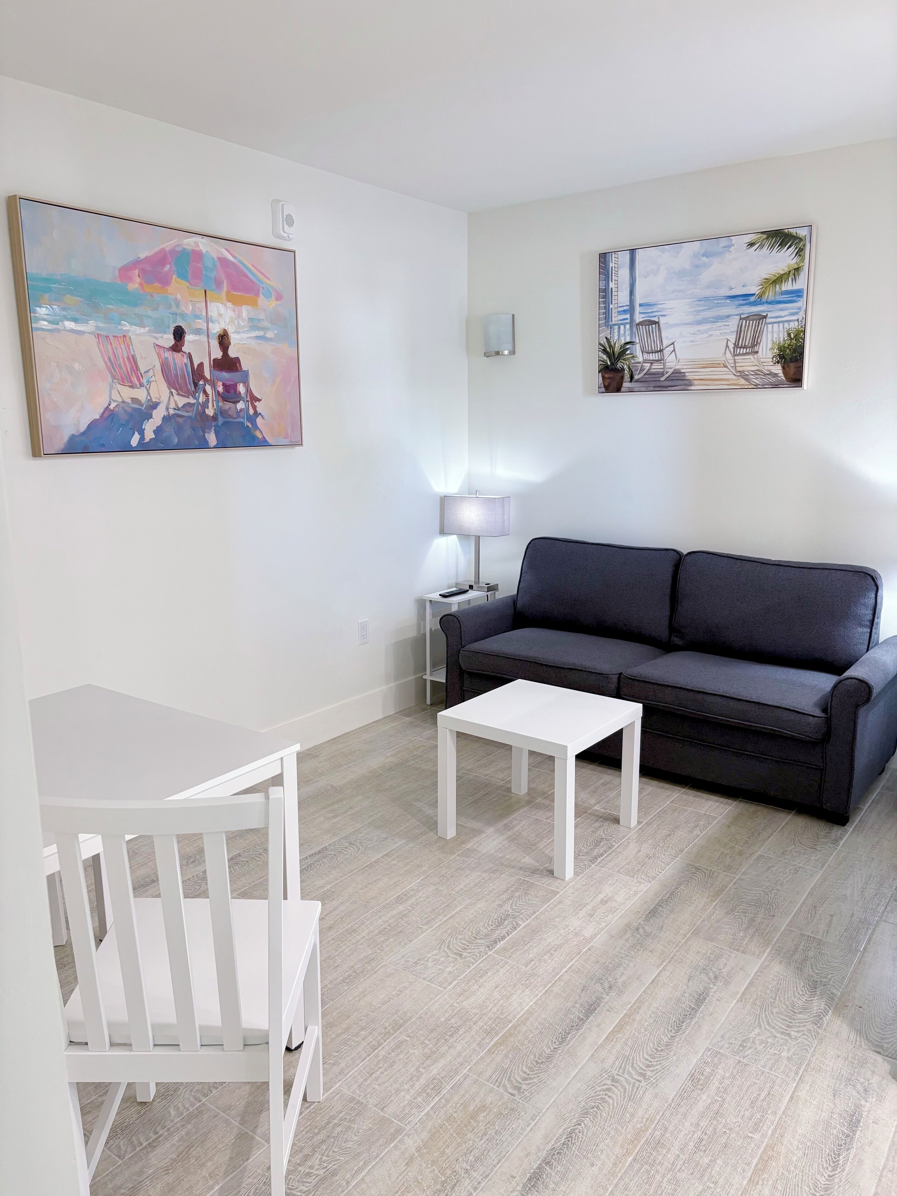 A living room with a dark grey sofa, a white coffee table, and two striped armchairs. There are two paintings on the walls, one depicting a beach scene and the other a balcony view.
