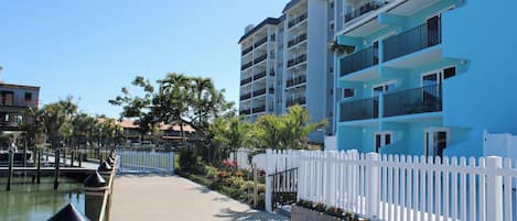 Un hotel frente al agua con un paseo marítimo de madera, cerca viva blanca y balcones con vistas al agua.