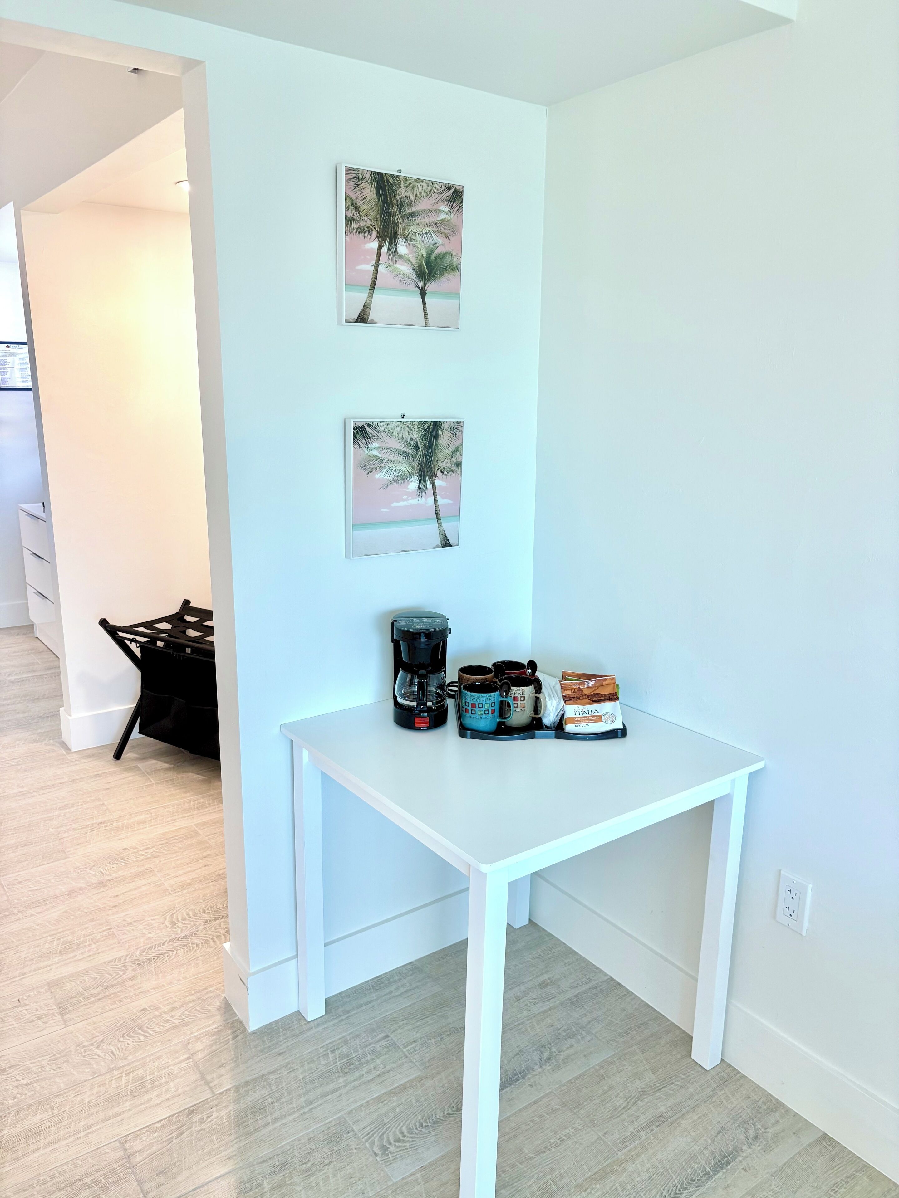 A small, white-framed table with a coffee maker, cups, and a bag of coffee in a bright room with light-colored flooring and white walls.