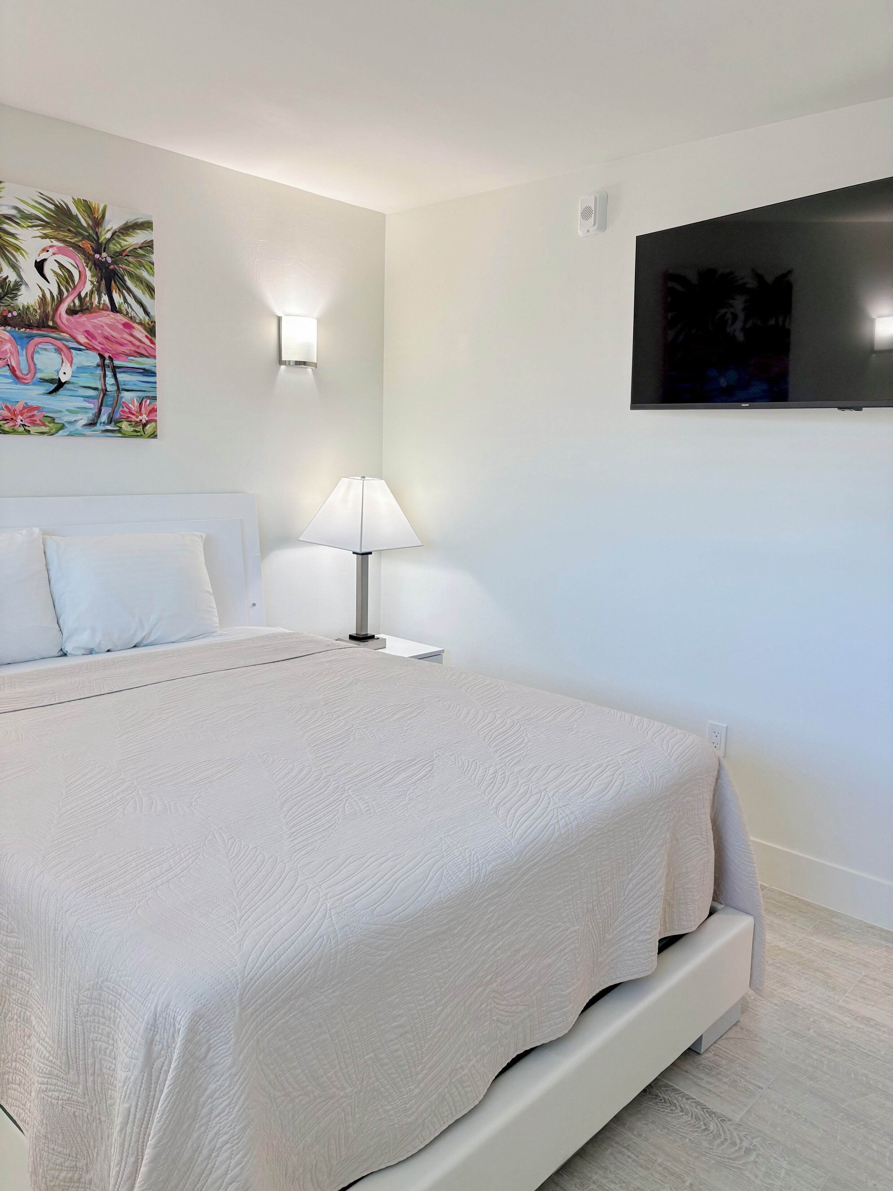 Un dormitorio con cama, televisor montado en la pared, mesita de noche con lámpara y un cuadro de flamencos.